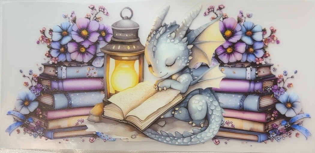 Sleepy blue dragon reading beside a glowing lantern, pastel flowers and stacked books fantasy tumbler wrap